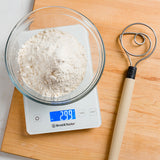 Bowl of flour on a baking scale alongside a dough whisk on a wooden cutting board.